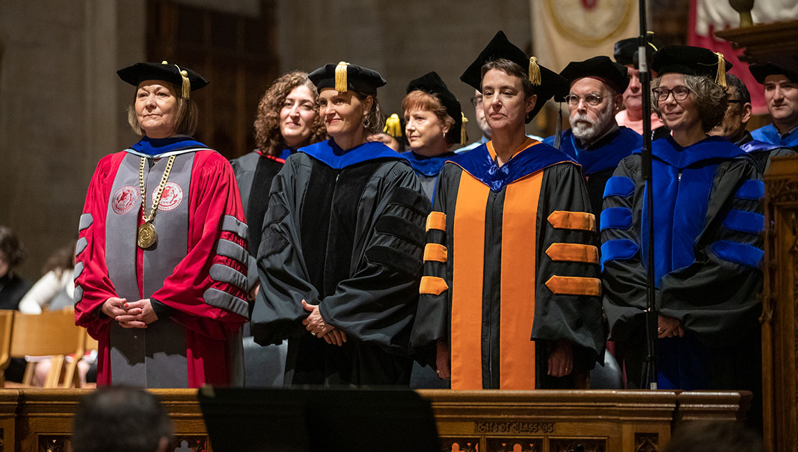 A group of adults dressed in academic robes stands inside a chapel ahead of a ceremony